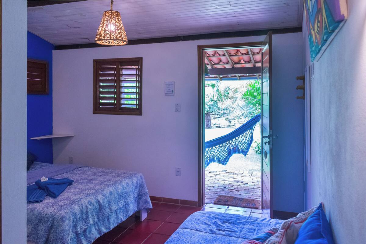 Ein Schlafzimmer mit zwei Betten, einem Fenster mit Rollläden und einer Tür zu einem Außenbereich mit einer Hängematte.