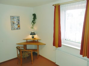 Ein Zimmer mit einem Eckschreibtisch, einem Stuhl, einer Lampe, einem Heizkörper und einem fenster mit Vorhängen.