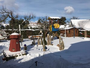A snow-covered outdoor area with a red lighthouse, a wooden play structure, and a small wooden building. - Haus Schumann - Ostsee - Halbinsel Darss - Ferienwohnung (Wieck a. Darß)