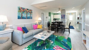 Ein Wohnzimmer mit einer Couch, einem Couchtisch und farbenfroher Kunst. Im Hintergrund ist ein Essbereich mit Stühlen und einem Tisch zu sehen.