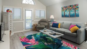 Ein Wohnzimmer mit grauem Sofa, bunten Kissen, einem tropischen Teppich, einem Gemälde und einem Fenster mit Jalousien.