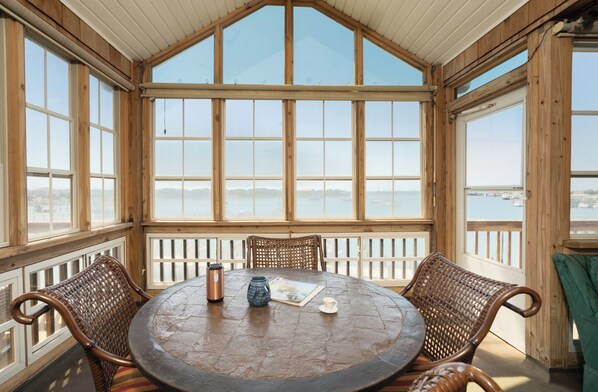 A wooden-paneled room with a large window overlooking a body of water, a round table with chairs, and a view of distant land.