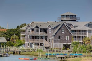A large, multi-story wooden house with a prominent tower, multiple balconies, and a dock with canoes.