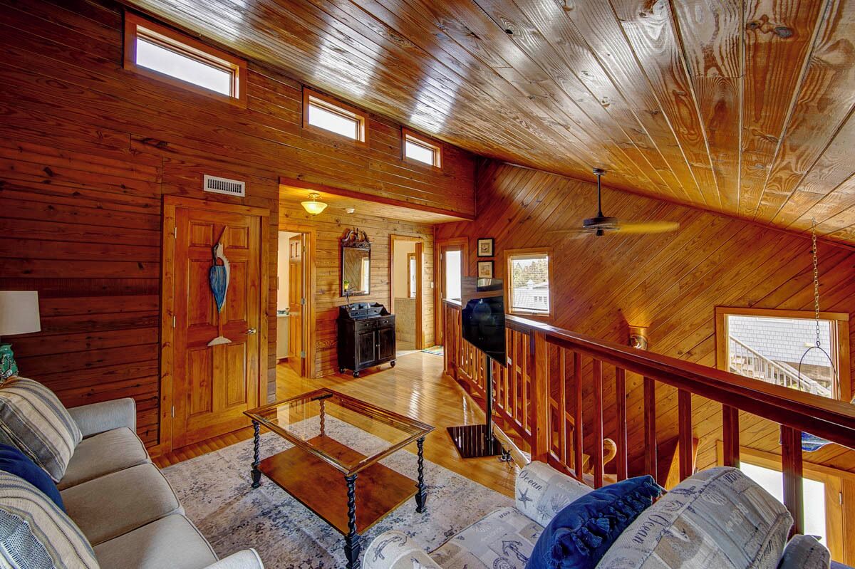 Il s’agit d’une pièce spacieuse de style « loft », avec des murs en bois et un plafond en pente. Elle comprend une grande fenêtre, un ventilateur de plafond et un coin salon avec un canapé et une table basse.