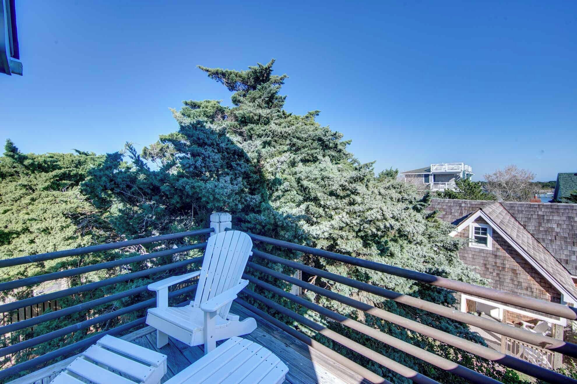 Una terraza de madera con una silla Adirondack blanca, rodeada de árboles y una casa al fondo.