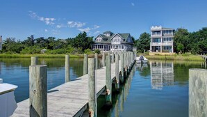 A waterfront property with a dock extending into a calm body of water, a large house with a balcony, and a smaller building adjacent to it.