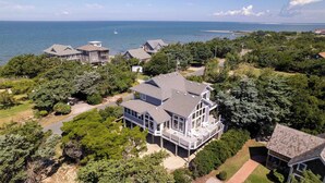 A large, multi-story house with a prominent balcony, surrounded by dense greenery and a view of the ocean.
