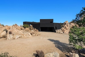 A modern building with a flat roof, surrounded by large rocks and sparse vegetation. - Black Desert House ft in Architectural Digest (Yucca Valley)