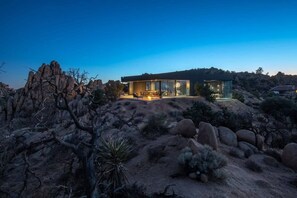 Une maison moderne dotée de grandes fenêtres, entourée d’un paysage désertique et d’un terrain rocheux.
