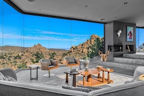 A modern living room with a large glass wall, a fireplace, and a view of rocky terrain. - Black Desert House ft in Architectural Digest (Yucca Valley)