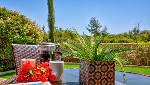 Una mesa con una cafetera, dos tazas, un helecho en maceta y flores rojas.