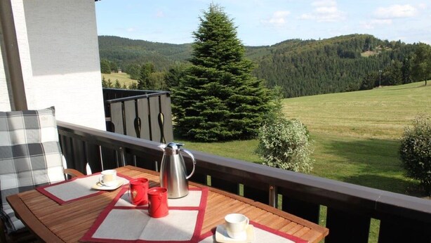 Ein Holztisch mit einer Metall-Kaffeekanne, zwei roten und zwei weißen Tassen auf einem Balkon mit Blick auf einen bewaldeten Hügel.