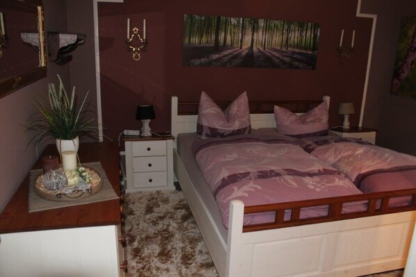 A bedroom with a bed, bedside tables, a nightstand, a wall-mounted lamp, a painting, and a potted plant. - Ferienwohnung Panorama Postwiese (Winterberg)