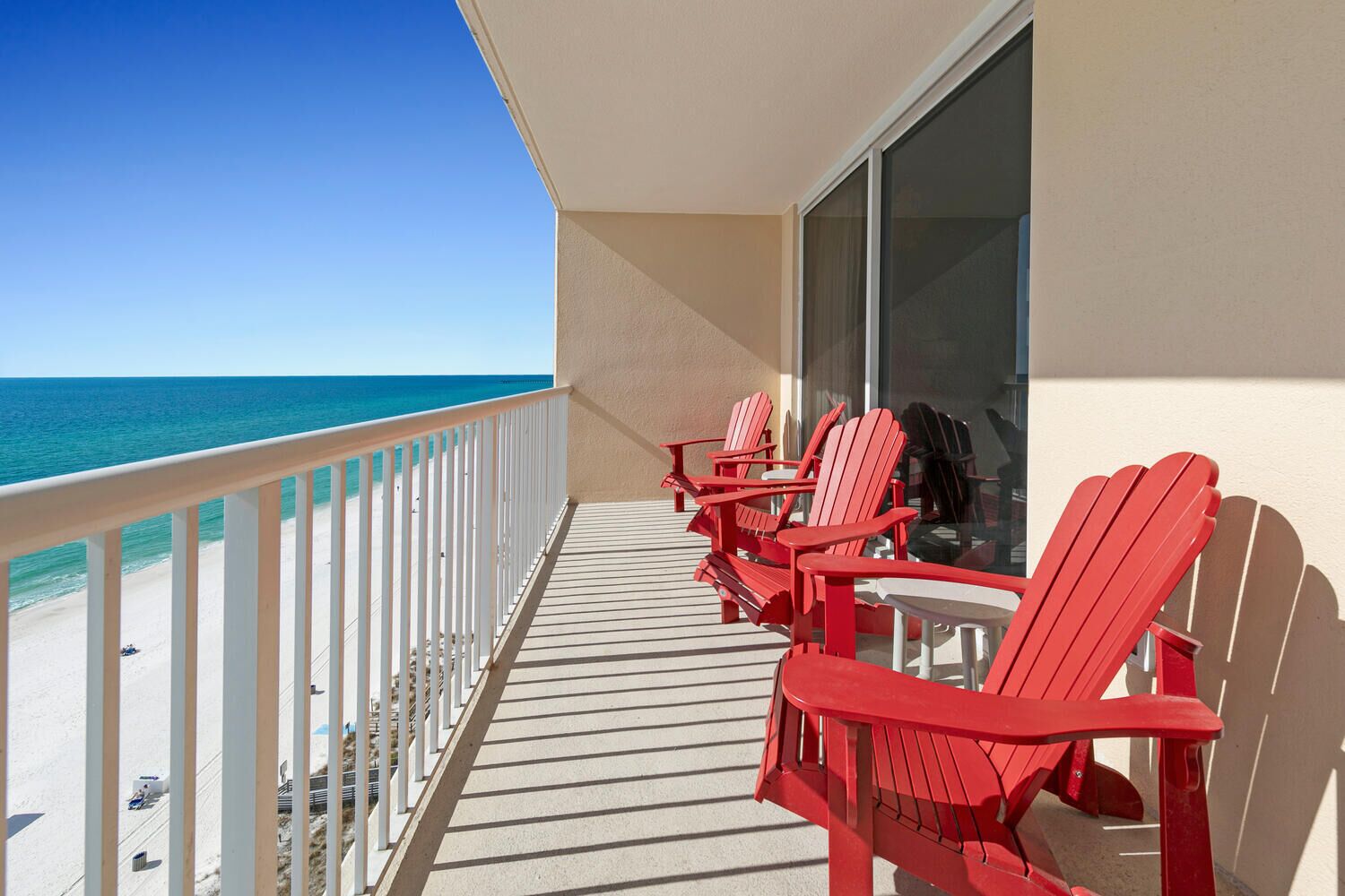 A balcony with a view of the ocean, featuring red Adirondack chairs and a glass door.