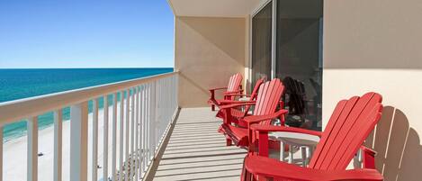 A balcony with a view of the ocean, featuring red Adirondack chairs and a glass door.