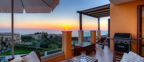A balcony with a view of the sunset, a grill, and outdoor furniture.