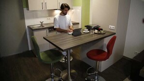 A modern kitchen with a person using a laptop, two bar stools, and a small dining table. - 3- Zimmer Apartment - Comfort Boardinghouse (Kornwestheim)