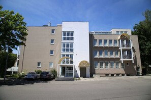A multi-story building with a central entrance, a parking area, and a small garden.
