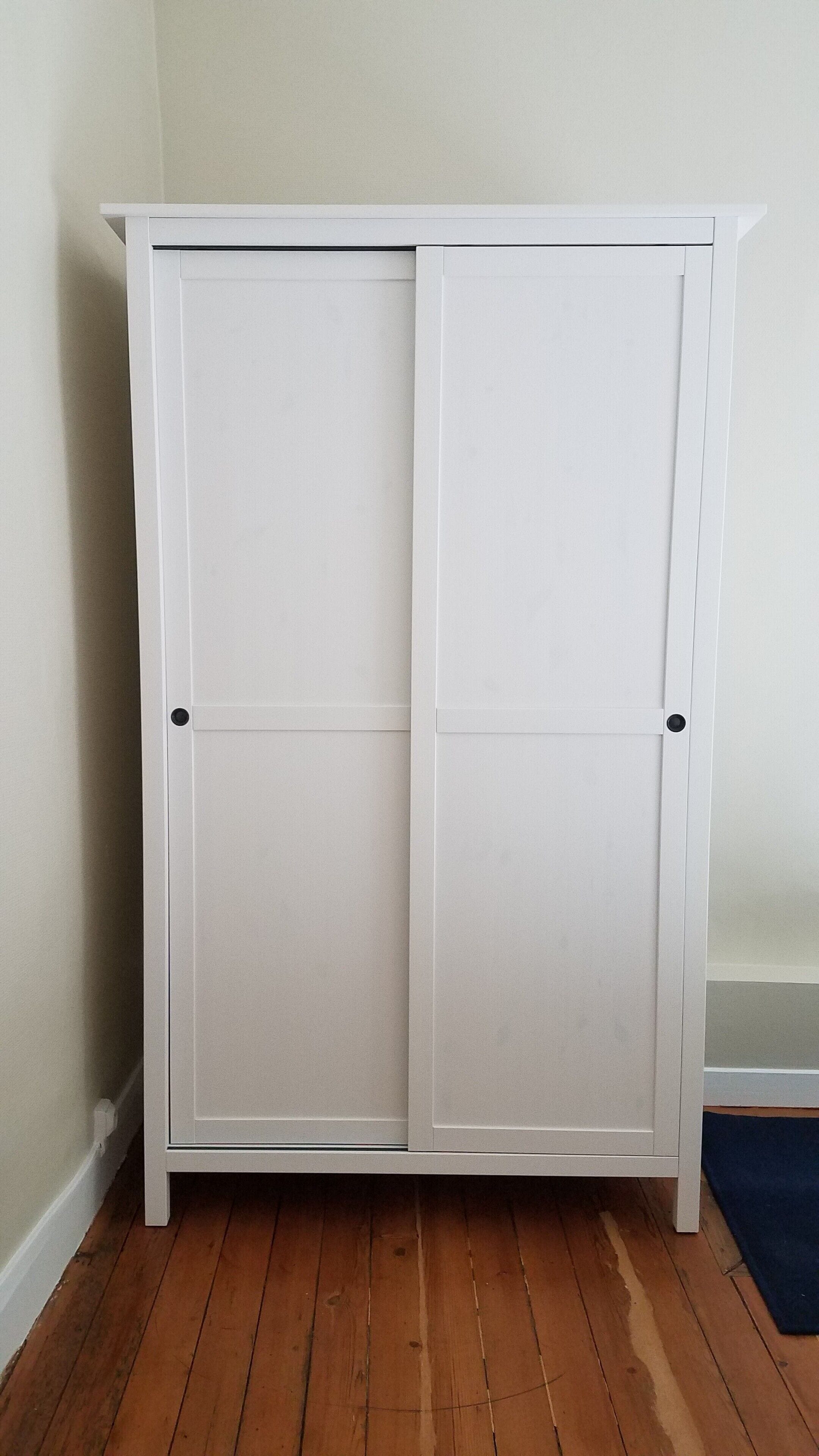 A white wardrobe with two doors and a flat top shelf, placed against a wall with a wooden floor.