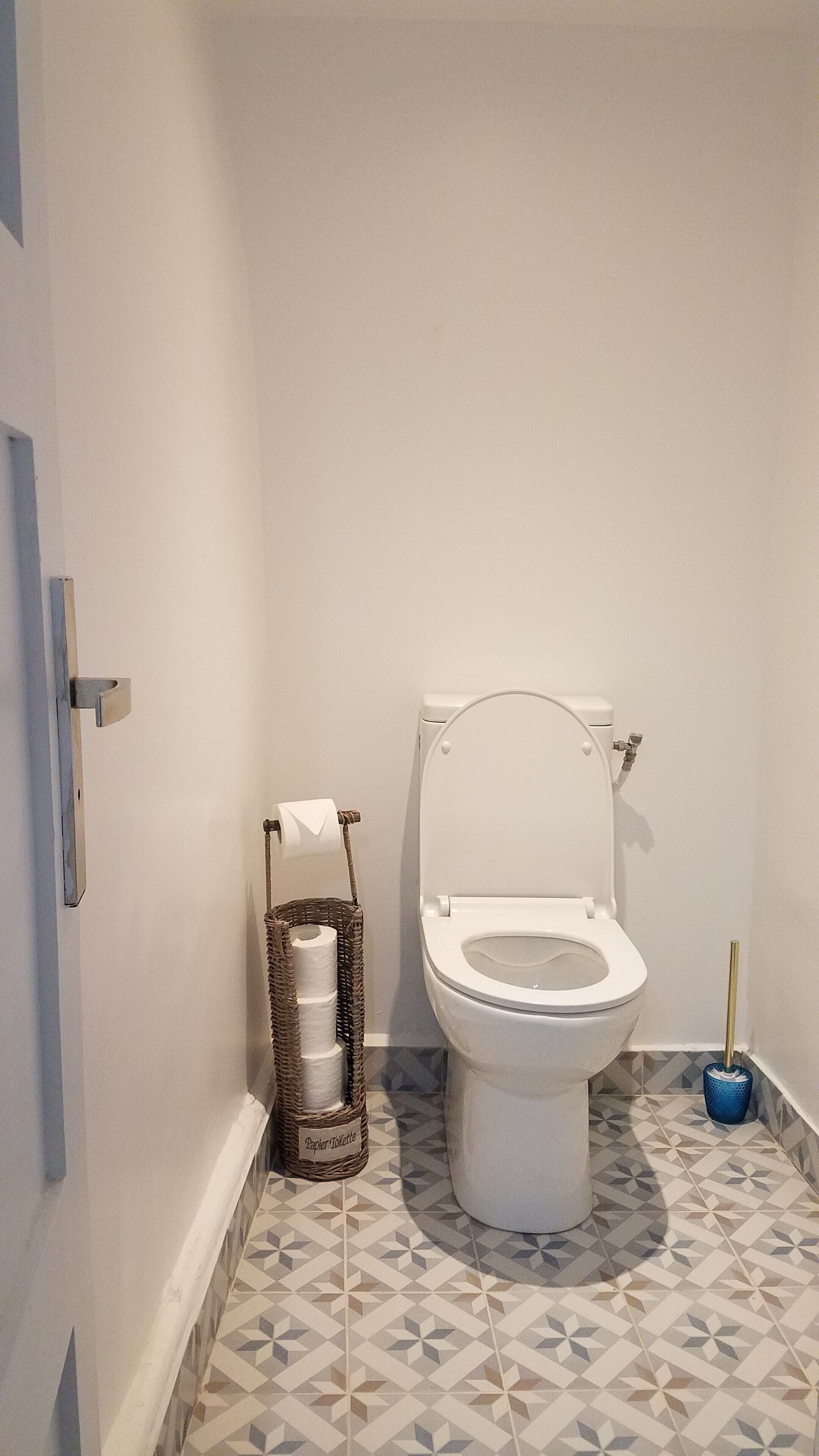 A small, clean bathroom with a toilet, a basket of toilet paper, and a blue toilet brush.