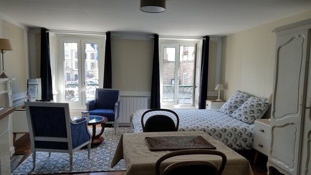 Quarto de hotel com cama, escrivaninha, cadeira, mesinha e vista da cidade através de grandes janelas.