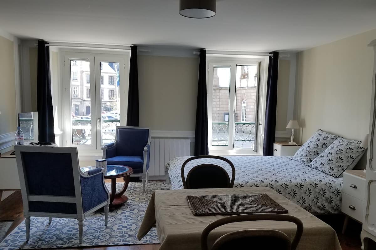 A hotel room with a bed, a desk, a chair, a small table, and a view of the city through large windows.