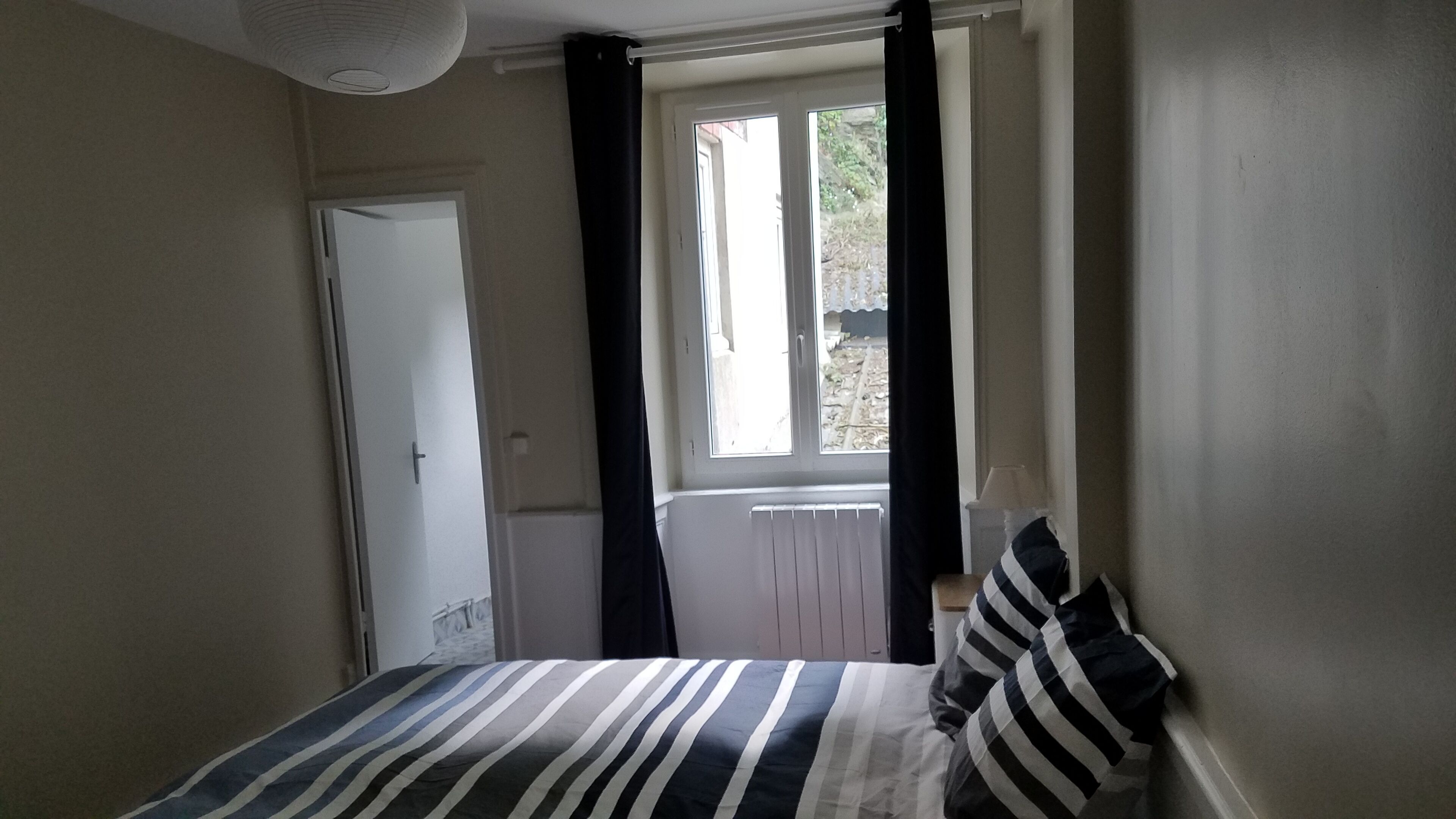 Un dormitorio con cama, ventana con cortinas, radiador y puerta.
