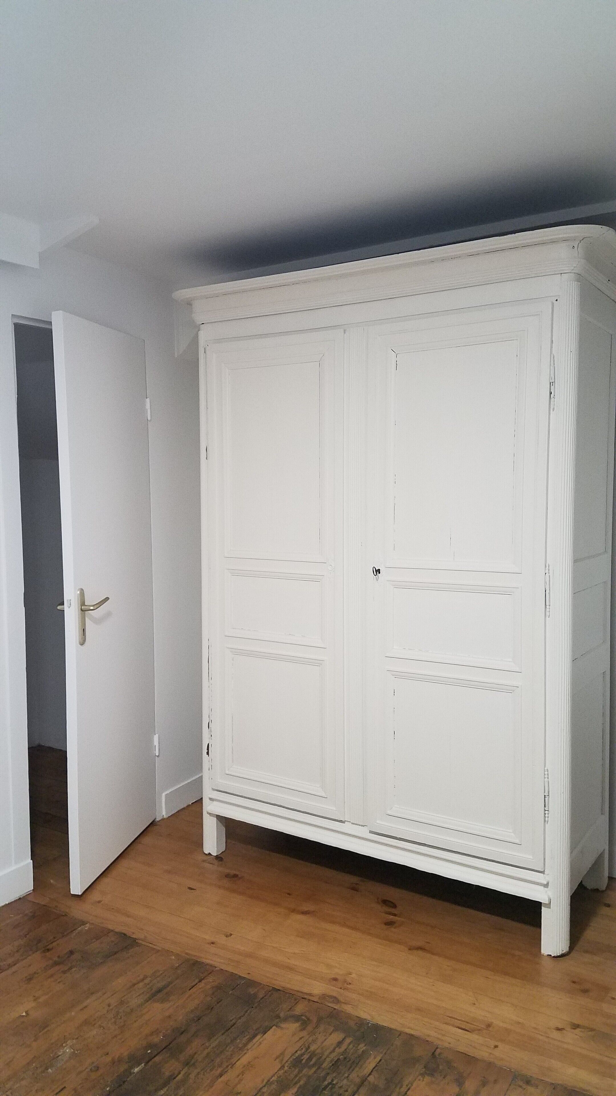 A white wardrobe with two doors and a small shelf, placed in a room with wooden flooring and a white door on the left.