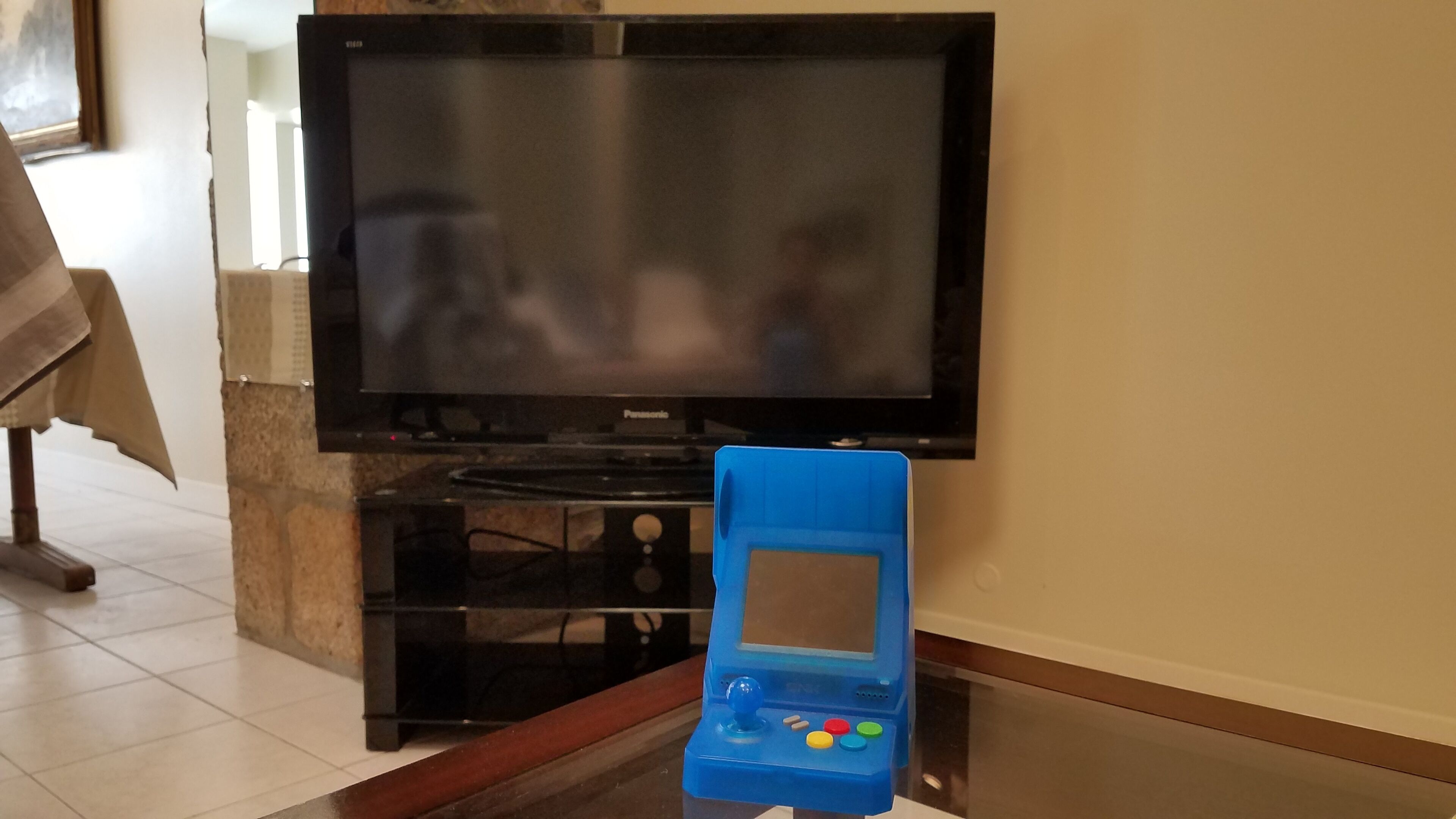 A living room with a flat-screen TV mounted on the wall, a blue gaming console on a glass table, and a brown table with a cloth.