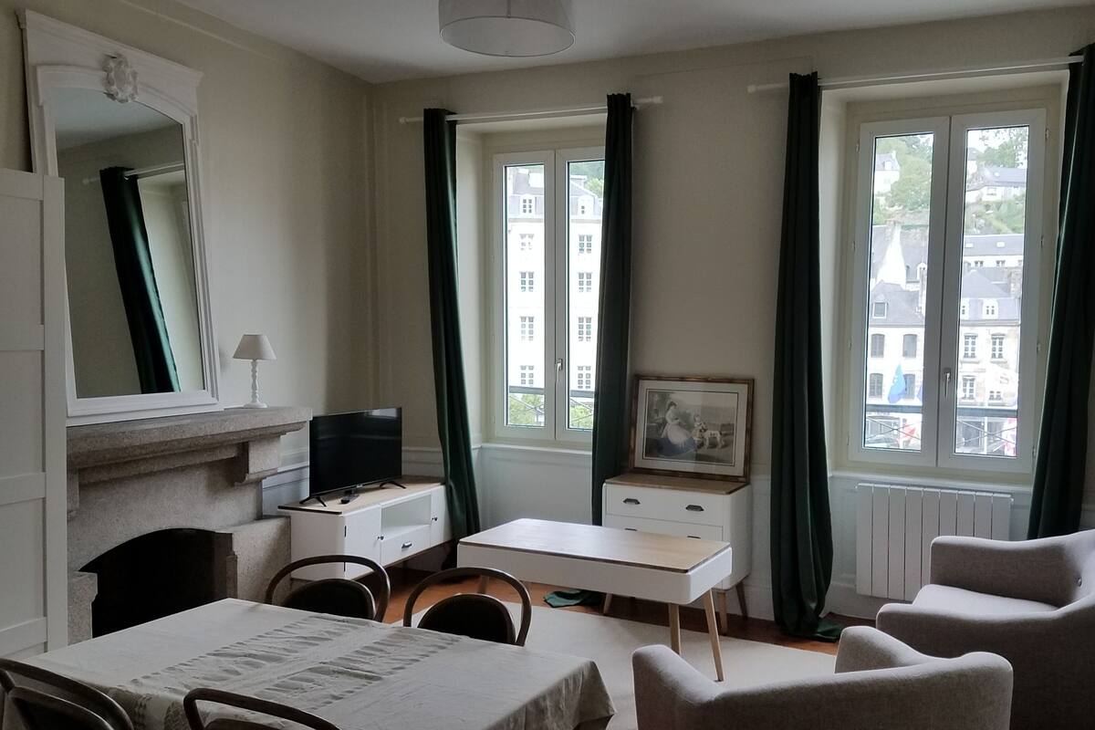A room with a dining table, chairs, a television on a stand, a small cabinet, a framed picture, and two windows with curtains.