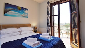 Un dormitorio con cama, almohadas, una mesita de noche, lámpara, un cuadro y vistas a través de una ventana.
