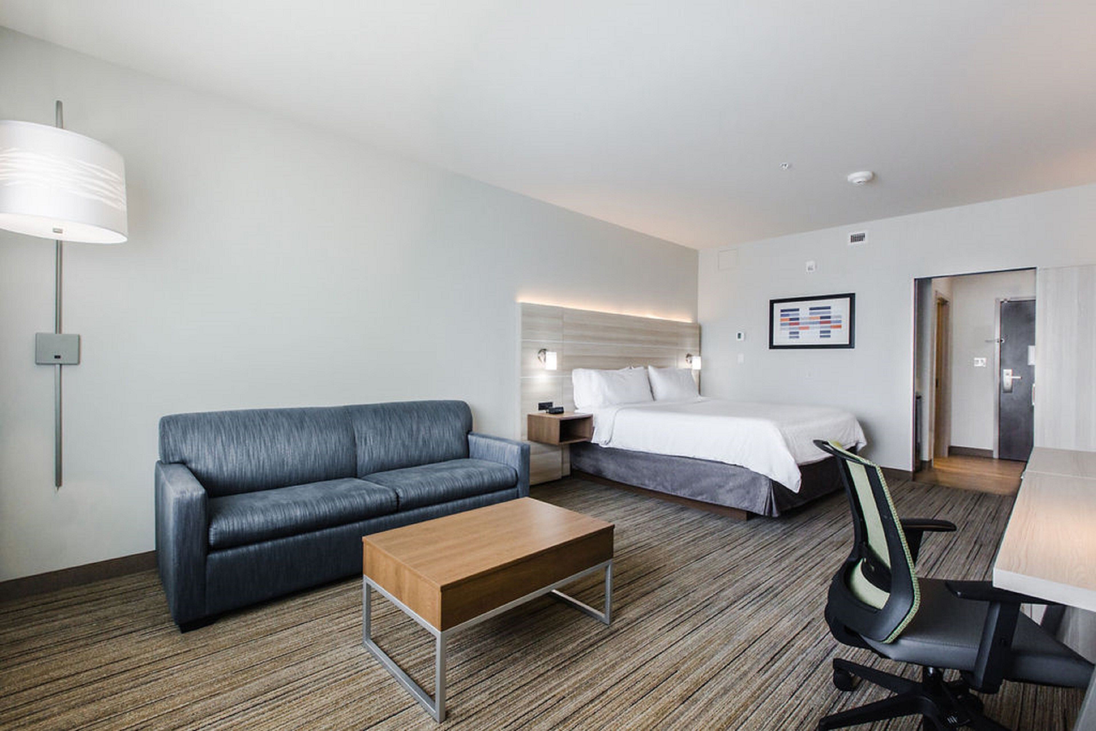 Una habitación de hotel con un sofá gris, una mesa de centro de madera, una cama con ropa blanca, un escritorio con una silla y una lámpara de pared.