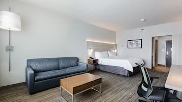 Una habitación de hotel con un sofá gris, una mesa de centro de madera, una cama con ropa blanca, un escritorio con una silla y una lámpara de pared.