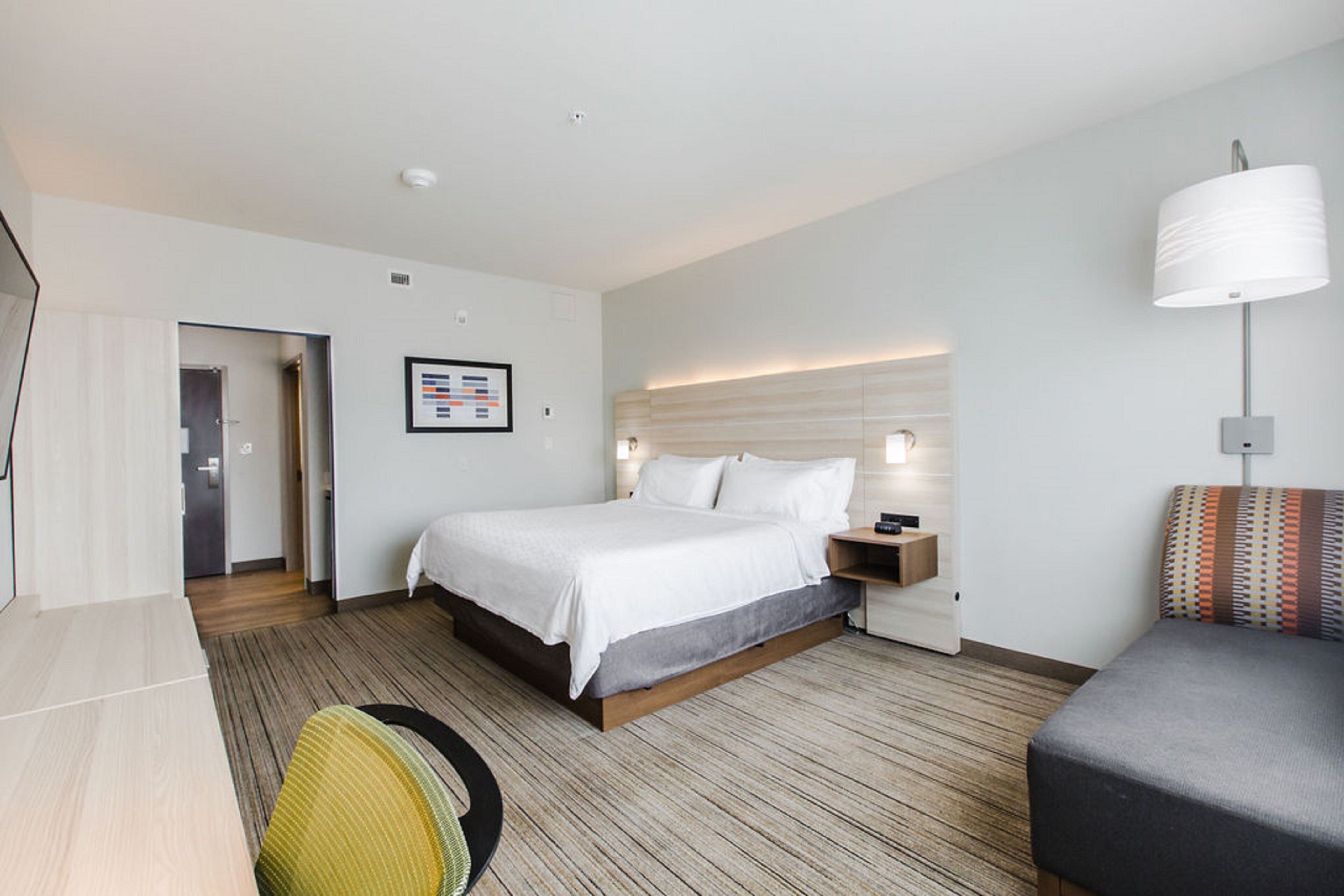 A modern hotel room with a large bed, bedside table, lamp, and a small seating area with a cushion.