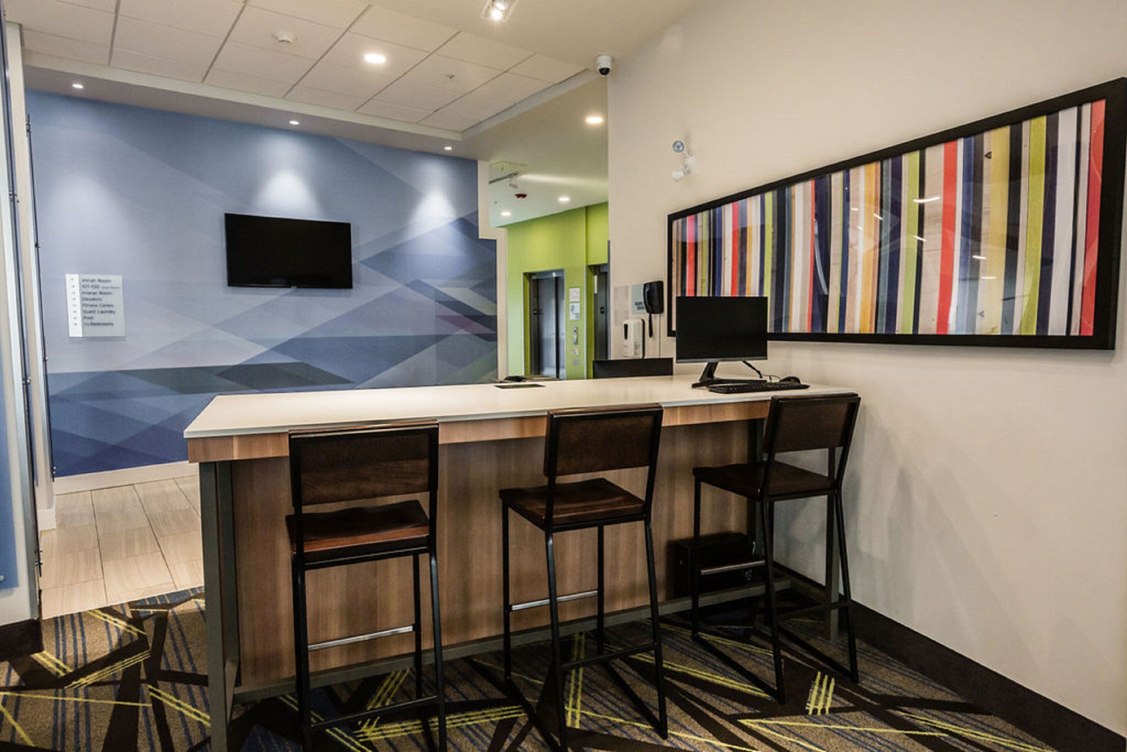 Un hall d’hôtel moderne avec un bar comprenant trois tabourets, un téléviseur à écran plat fixé au mur et une œuvre d’art colorée à rayures au-dessus du bar.