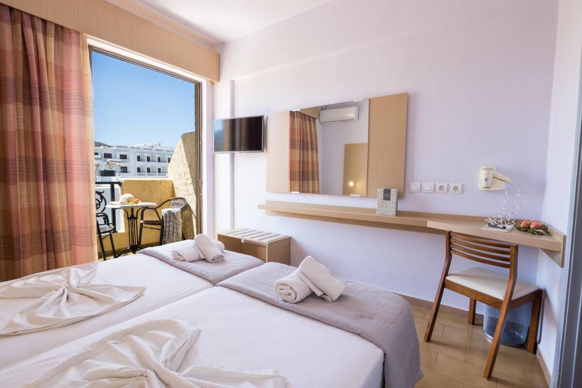A hotel room with a large bed, a desk, a chair, a television, and a balcony with a view of buildings.