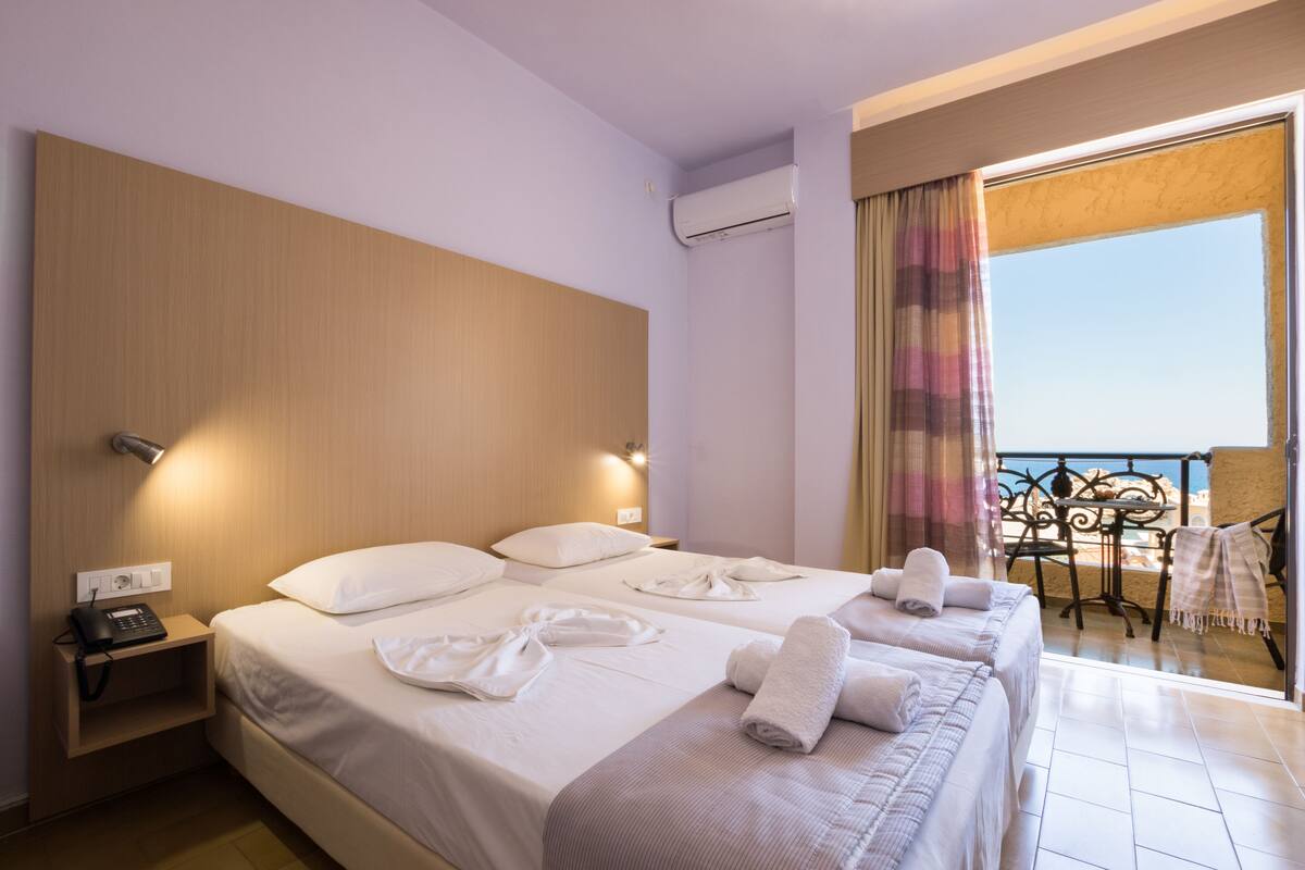 A hotel room with two beds, a bedside table with a phone, an air conditioning unit, and a balcony with a view.