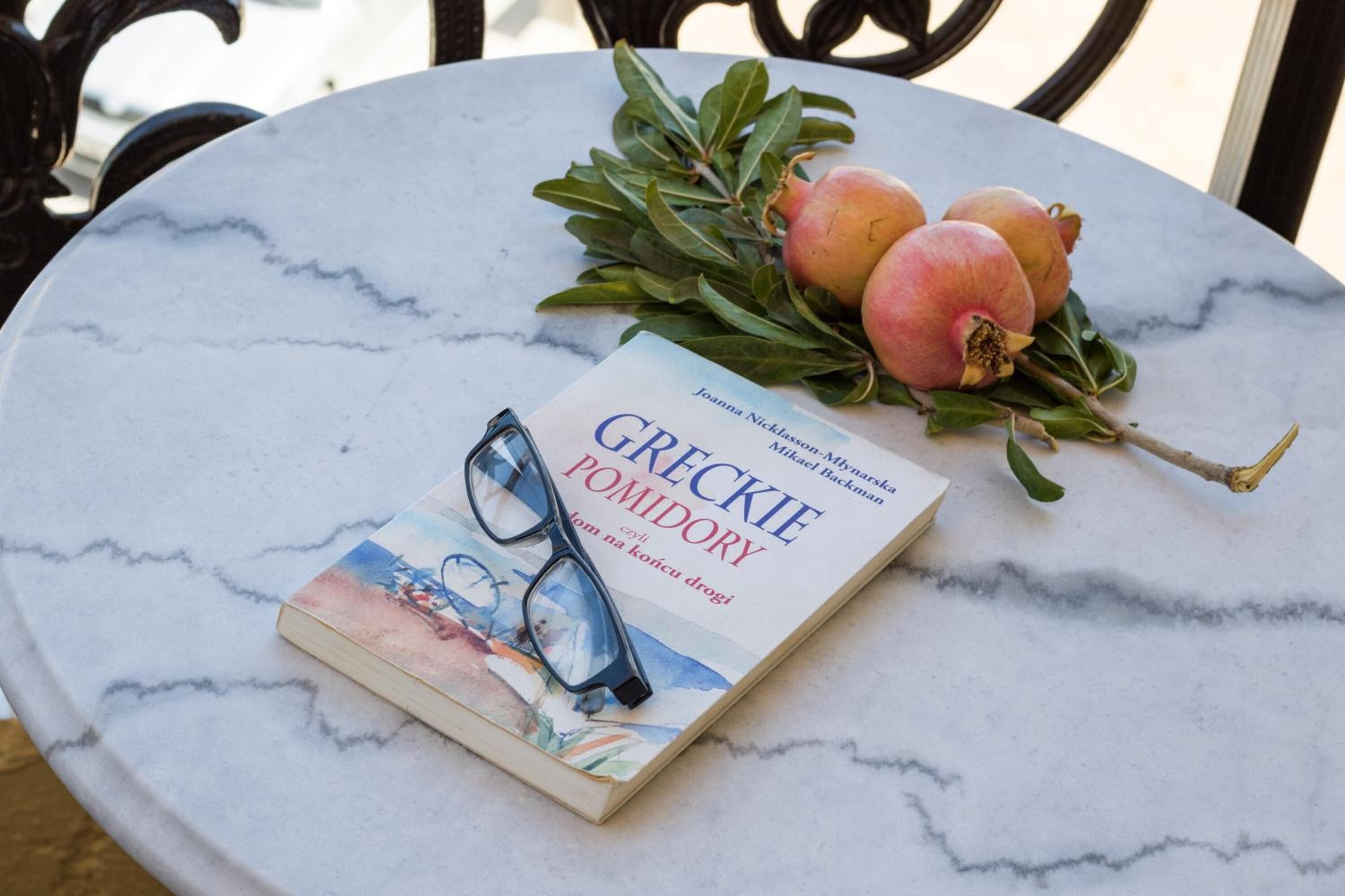 Ein Buch mit dem Titel 'GRECKIE POMIDORY', zusammen mit Brillen und Granatapfelkernen auf einer Marmortafel.
