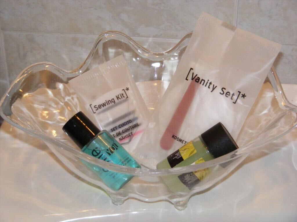 A clear glass bowl containing a sewing kit, a vanity set, and various cosmetic bottles.