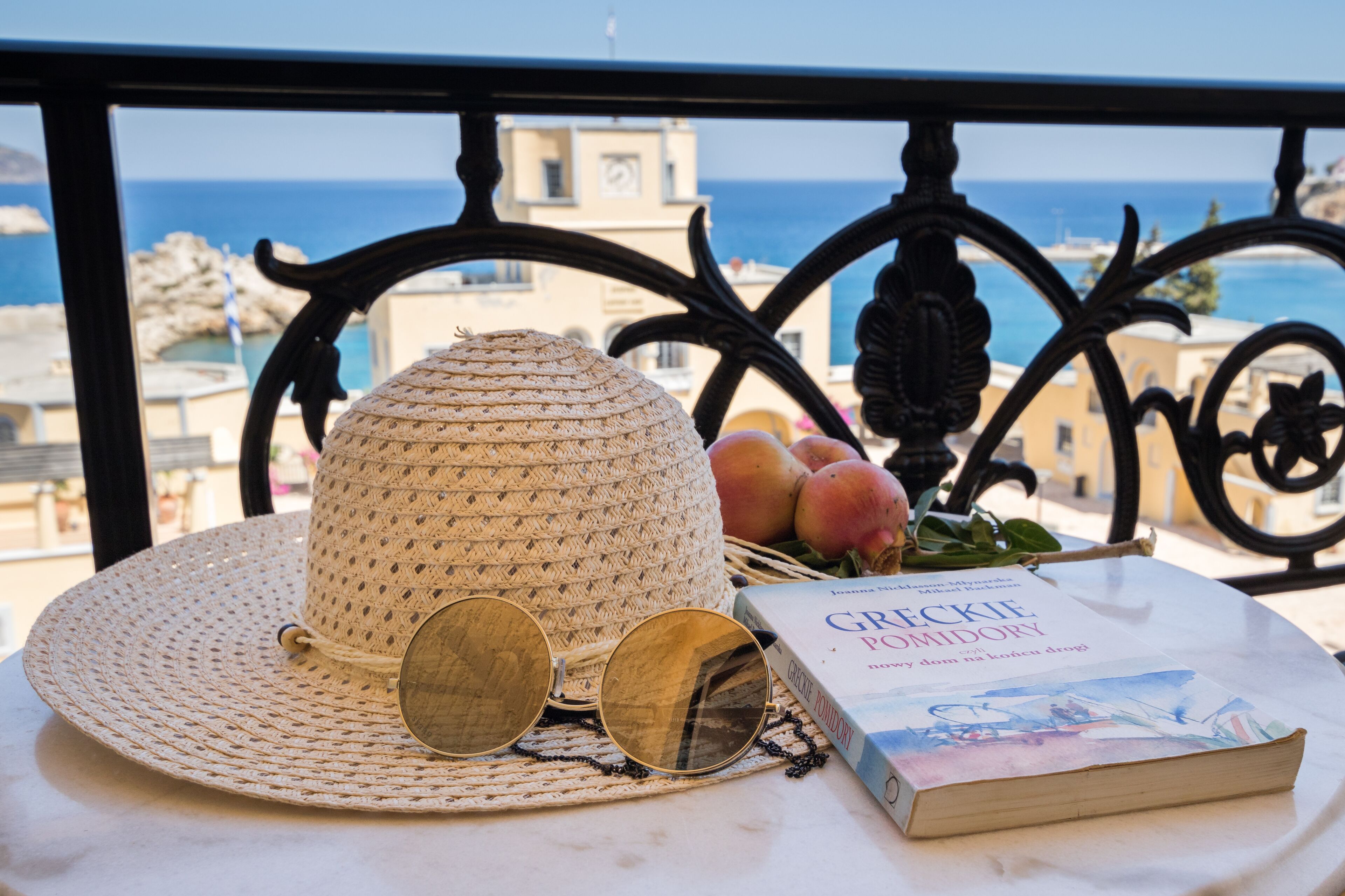 Een strohoed, zonnebril, een boek en een mand met fruit op een balkon met uitzicht op zee en gebouwen.