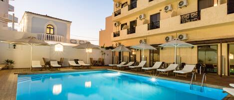 A swimming pool with lounge chairs and umbrellas, surrounded by a building with balconies and air conditioning units.