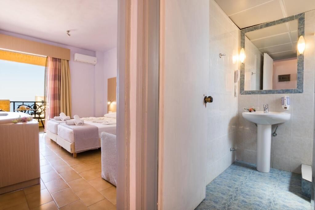 Habitación de hotel con cama, zona de estar con mesa y sillas, baño con lavabo y espejo, y vistas al exterior.