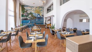 A spacious dining area with multiple tables and chairs, a mural on the wall, and a high ceiling with recessed lighting. - Atlantis Hotel (Karpathos)