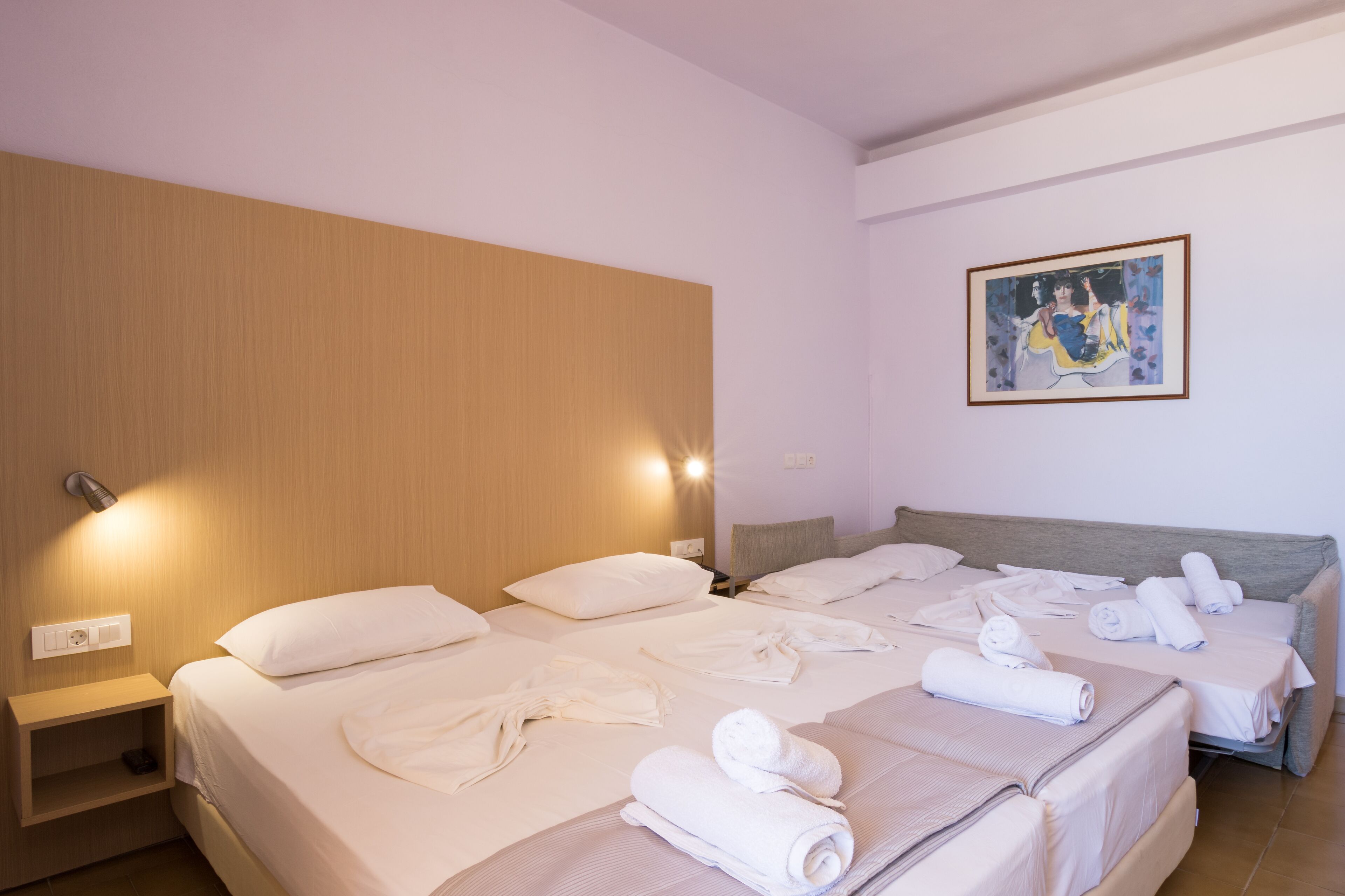 Habitación de hotel con dos camas, cada una con ropa blanca y toallas. Se observa un cabecero de madera en el lado izquierdo, un cuadro en la pared y una mesita de noche con una lámpara.