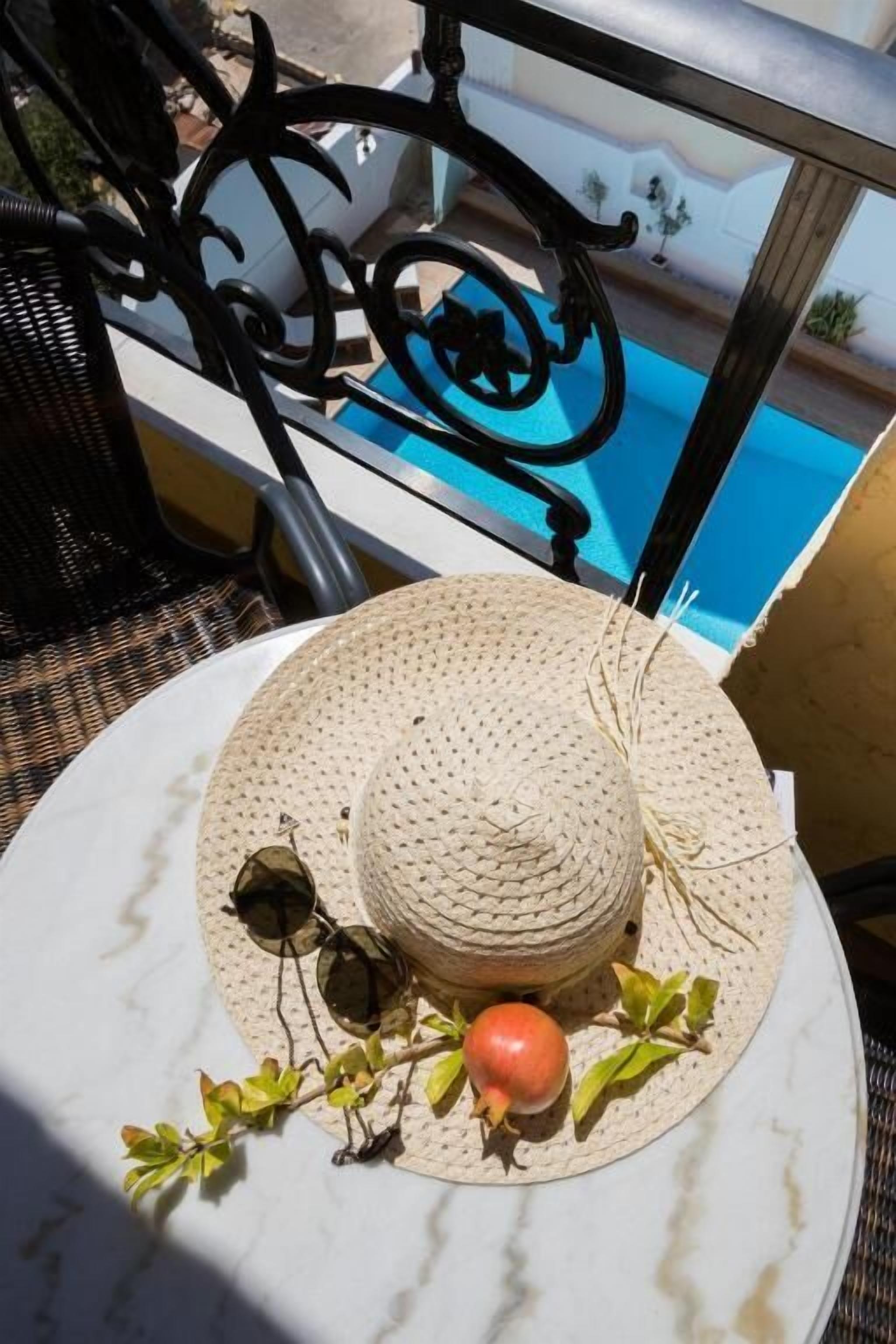 A round table with a woven hat, sunglasses, and a pomegranate on a balcony overlooking a swimming pool.