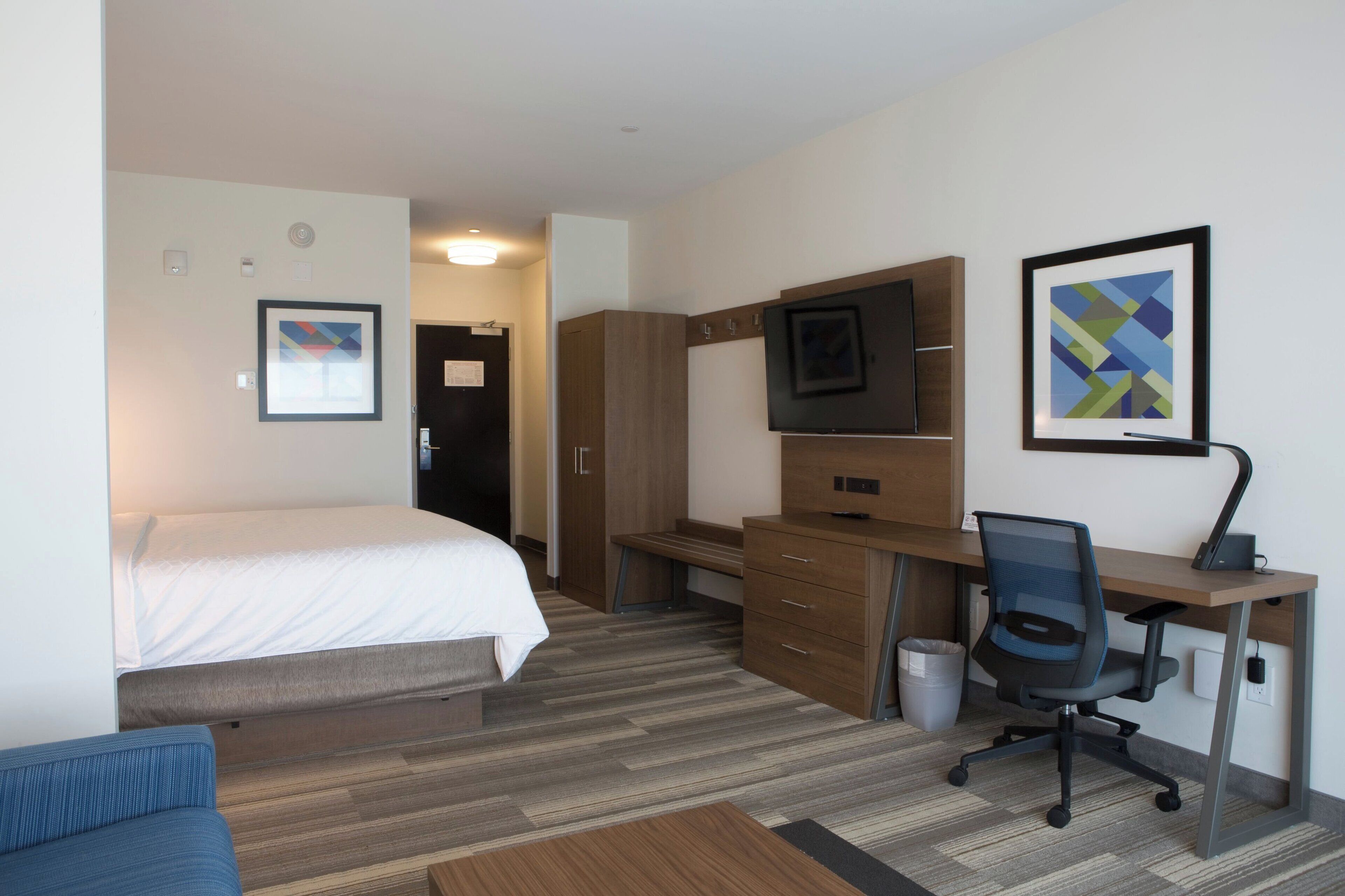 A hotel room with a bed, a desk with a chair, a TV, and artwork on the walls.
