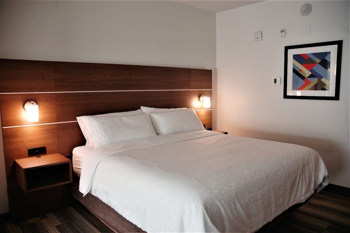 Un lit bien fait, avec du linge de lit blanc, une tête de lit en bois, des lampes de chevet et une œuvre d’art abstraite encadrée accrochée au mur.
