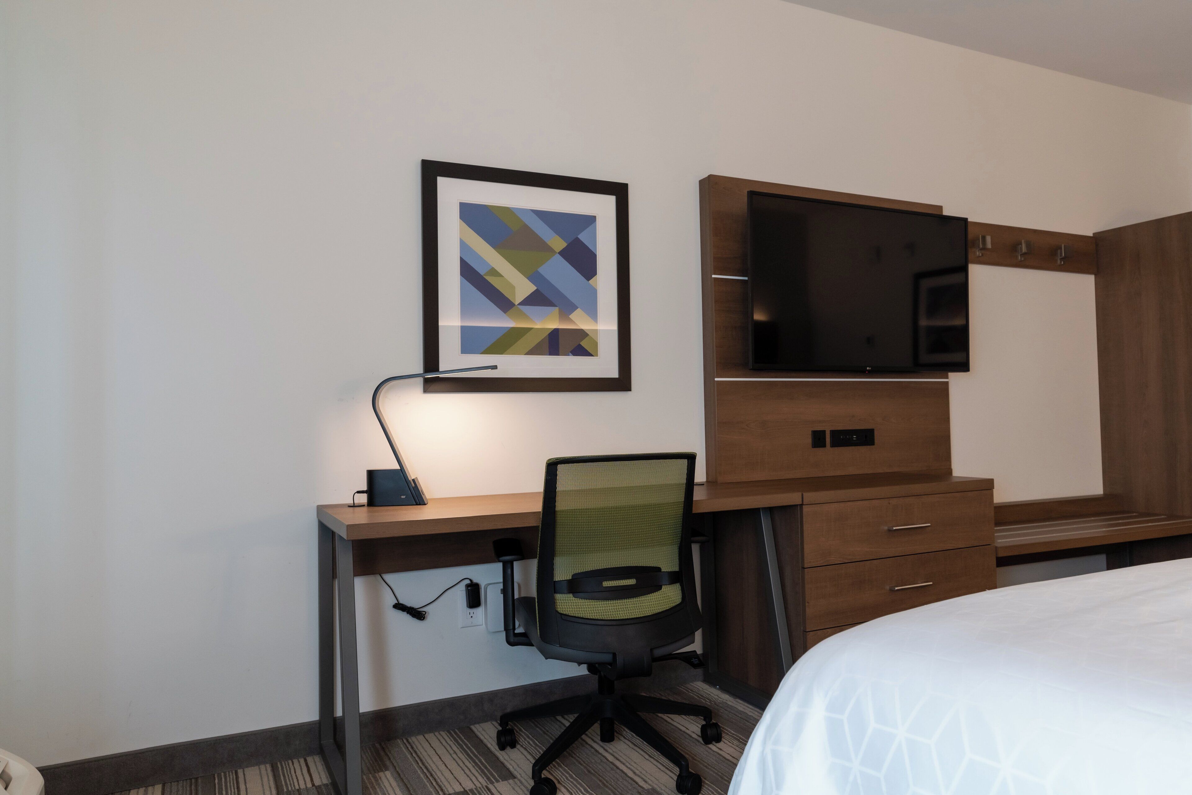 A hotel room with a bed, a desk with a chair, a flat-screen TV, a framed picture on the wall, and a wooden cabinet.