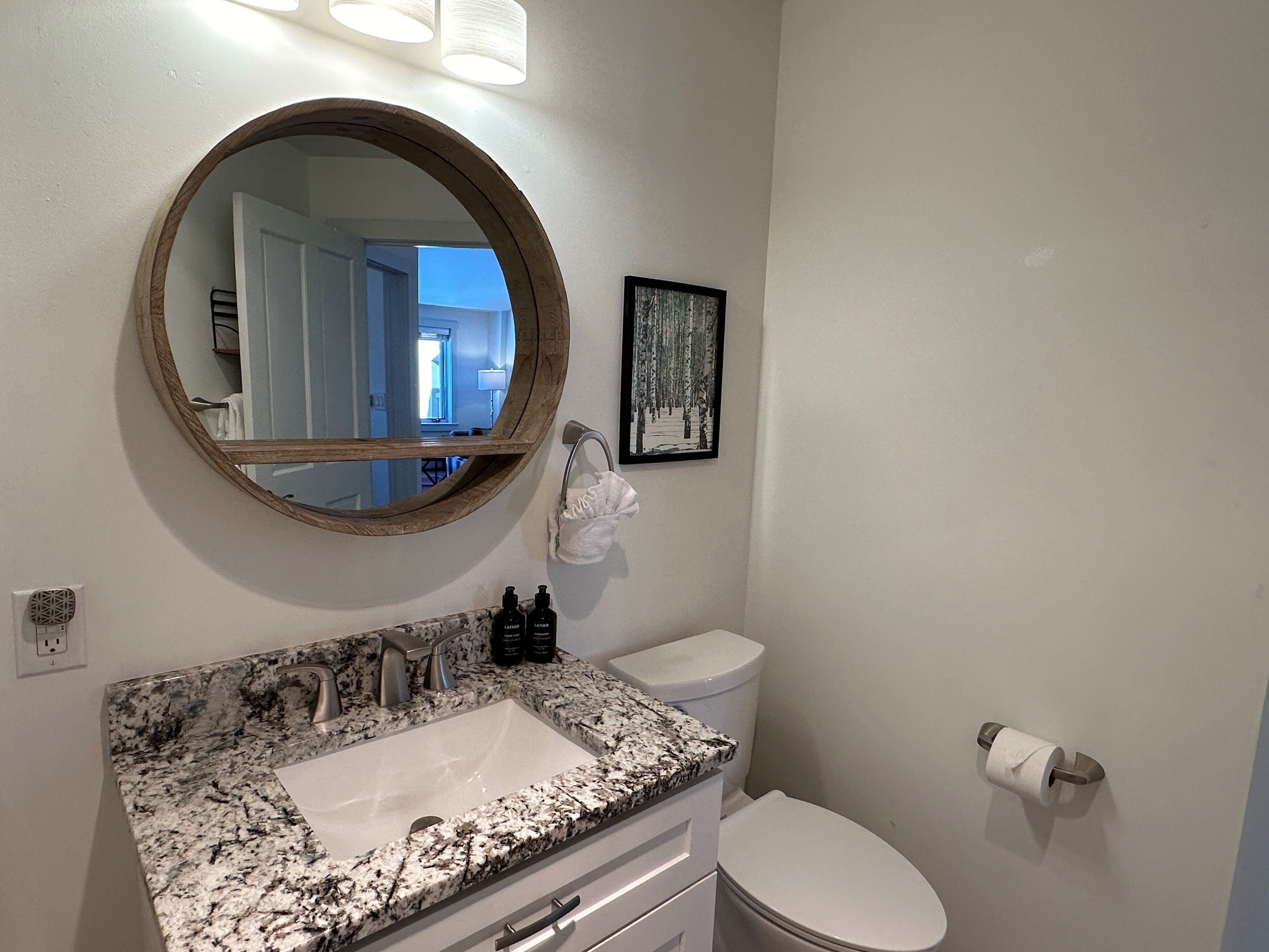 Une salle de bain avec un lavabo à surface en granit, des toilettes, un miroir rond et un tableau encadré au mur.