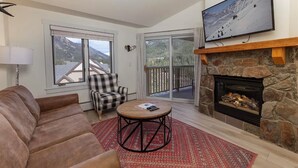 A living room with a brown leather sofa, a plaid armchair, a round coffee table, a fireplace, and a large TV mounted on the wall.